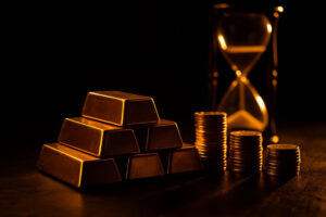 Pila di lingotti d’oro identici disposti a piramide con monete accanto, sullo sfondo una clessidra sfocata che rappresenta il passare del tempo e il valore crescente dell’oro.